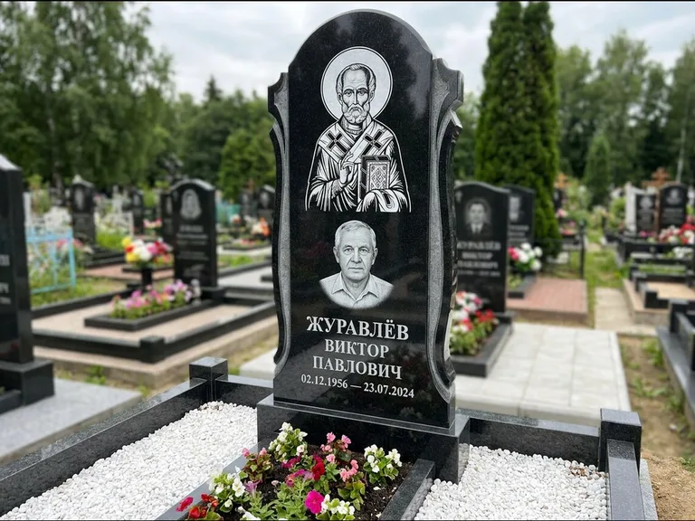 Памятник с гравировкой иконы: стела с образом в полный рост на обороте — гравировка образа Матроны Московской, низкие кусты спиреи в цвету, работа «Лебедева» в Липецке
